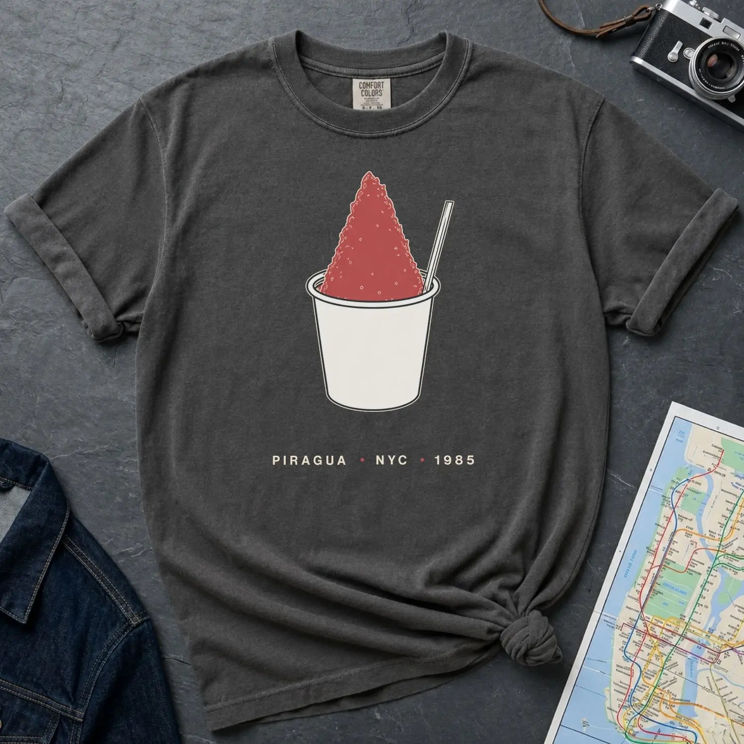 Pepper colored Comfort Colors t-shirt with a piragua graphic and text, placed on a dark surface with a map and denim jacket.