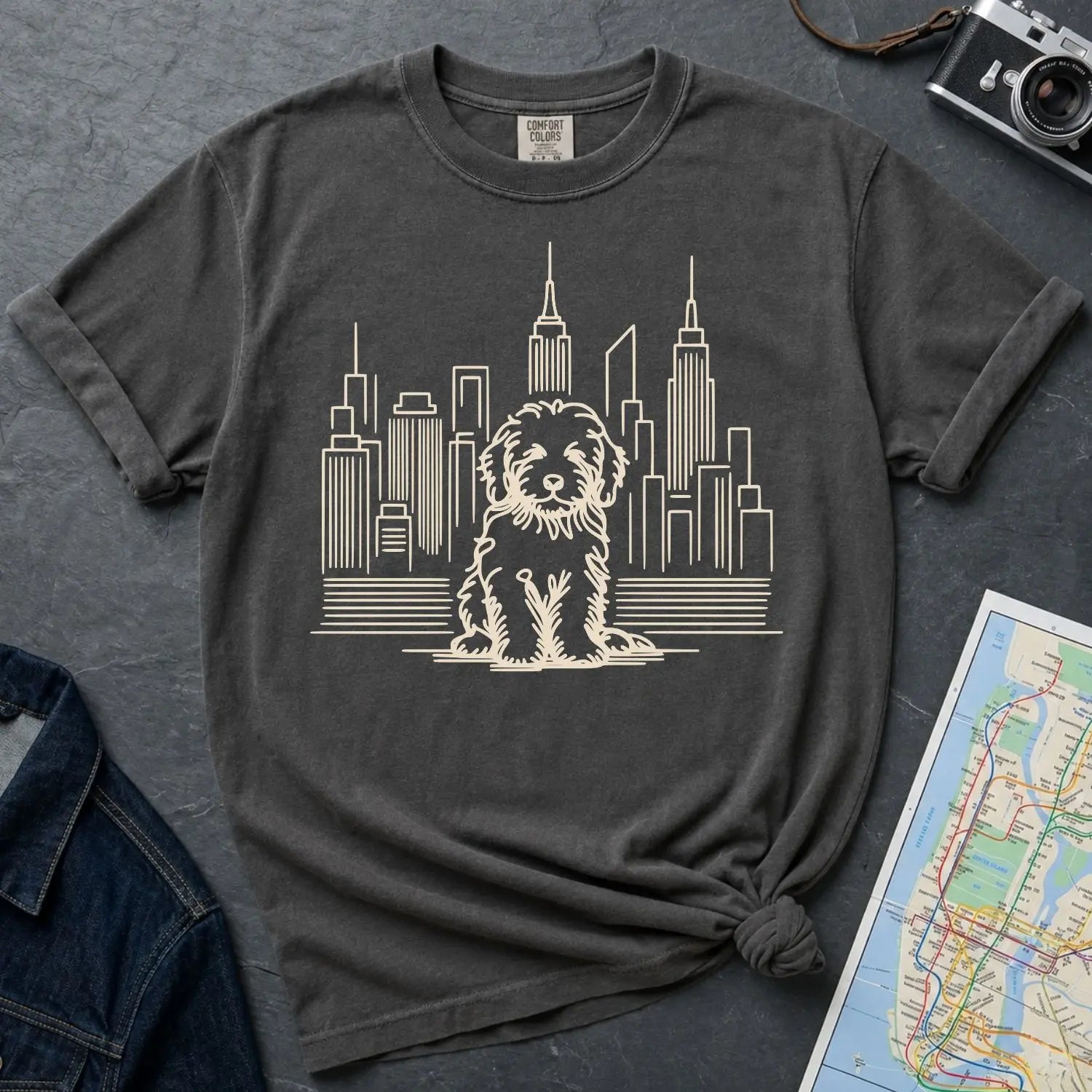Pepper colored Comfort Colors t-shirt with a Golden Doodle and cityscape graphic on a dark surface with a denim jacket and a map.
