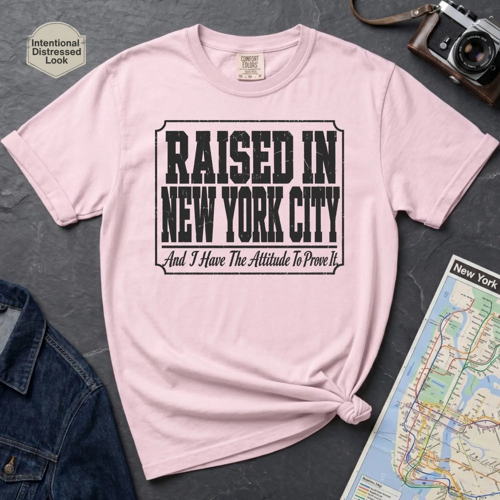 Pink t-shirt with 'Raised in New York City' text on a dark surface with a jean jacket and a map.