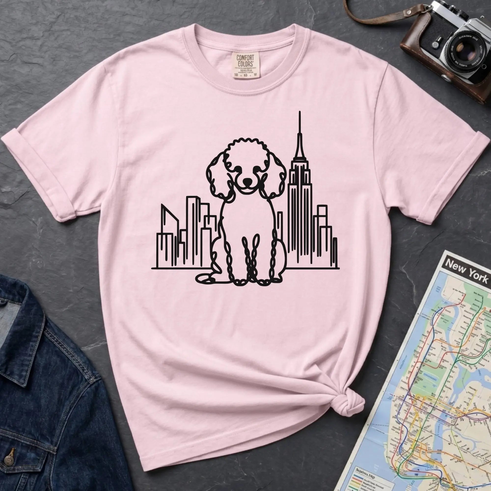 Pink t-shirt with a poodle and cityscape design on a dark surface with a map and camera.