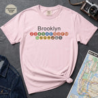 Pink t-shirt with 'Brooklyn' and subway line numbers on a dark surface with a map and camera.