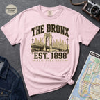 Pink t-shirt with 'The Bronx Est. 1898' design on a dark surface with a map and camera.