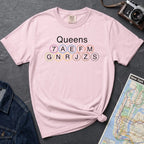 Pink t-shirt with 'Queens' and subway line numbers on a dark surface with a map and camera.