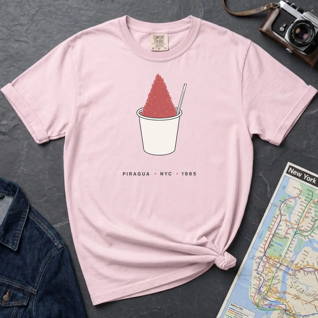 Pink Comfort Colors t-shirt with a piragua graphic and text, placed on a dark surface with a map and denim jacket.