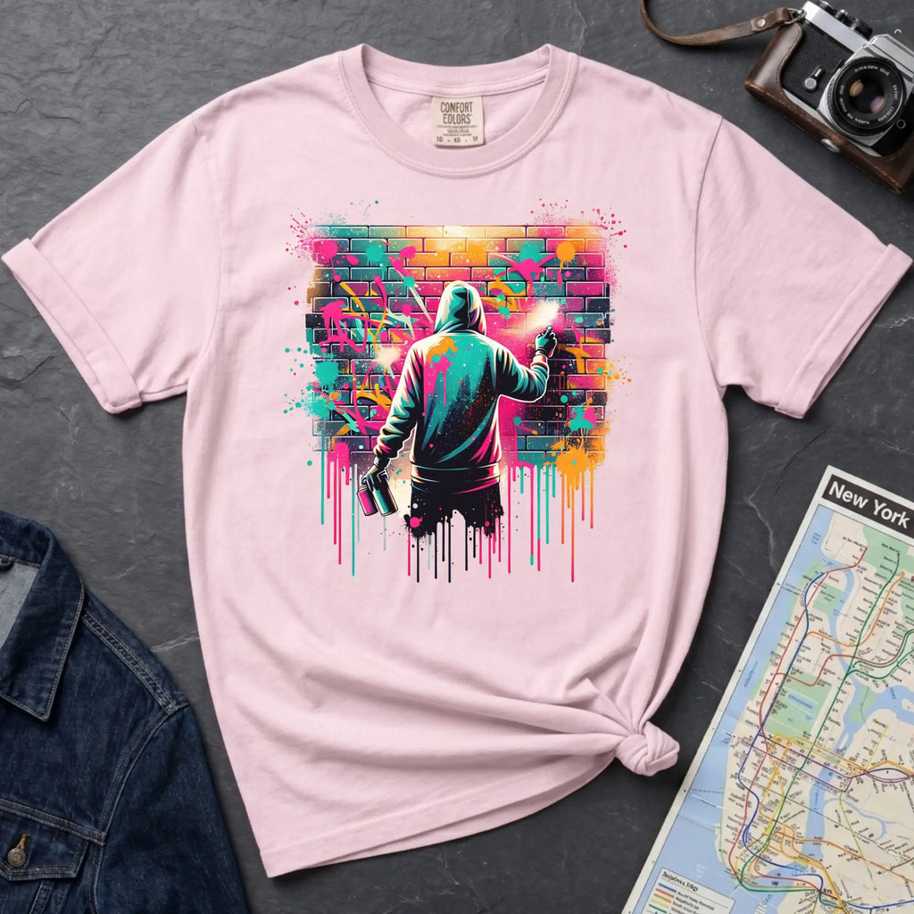 Pink t-shirt with a graphic of a person spray painting a colorful mural on a brick wall.