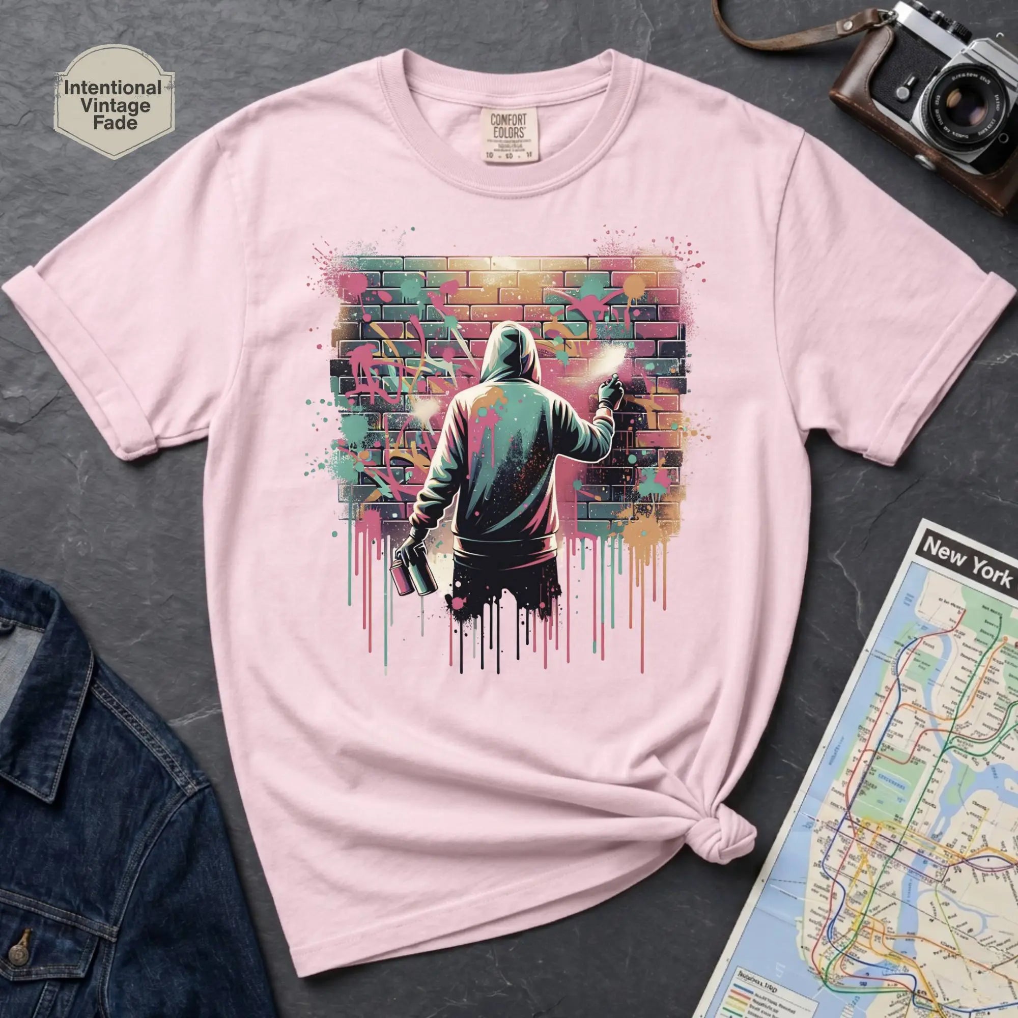 Pink t-shirt with a graphic design of a person spray painting a colorful wall, placed on a dark surface with a map and camera.