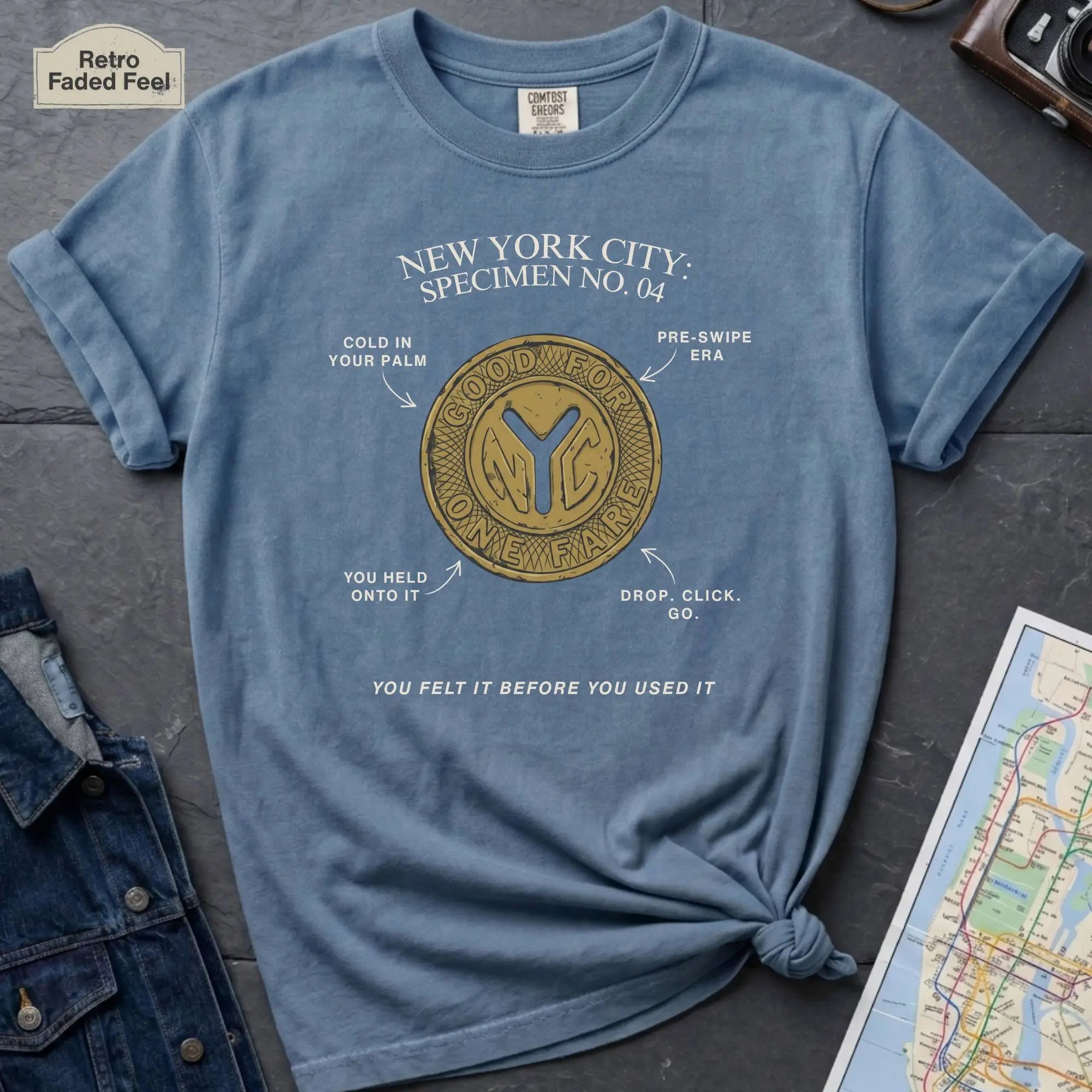 Blue t-shirt with New York City token design on a gray surface with a map and camera.