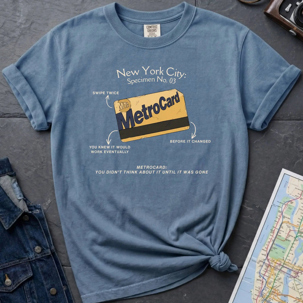 Blue t-shirt with New York City MetroCard graphic on a gray surface with a map and jacket.