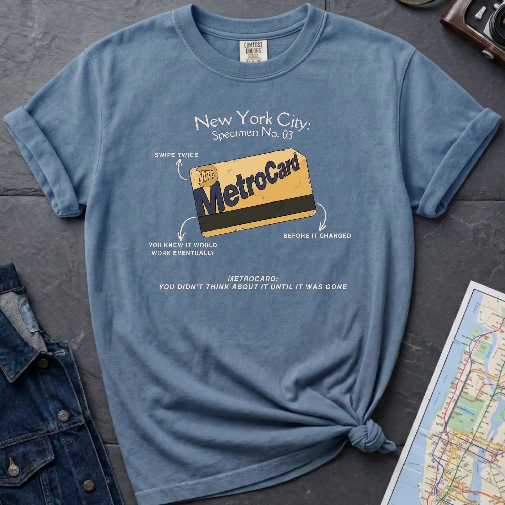 Blue t-shirt with New York City MetroCard graphic on a gray surface with a map and jacket.
