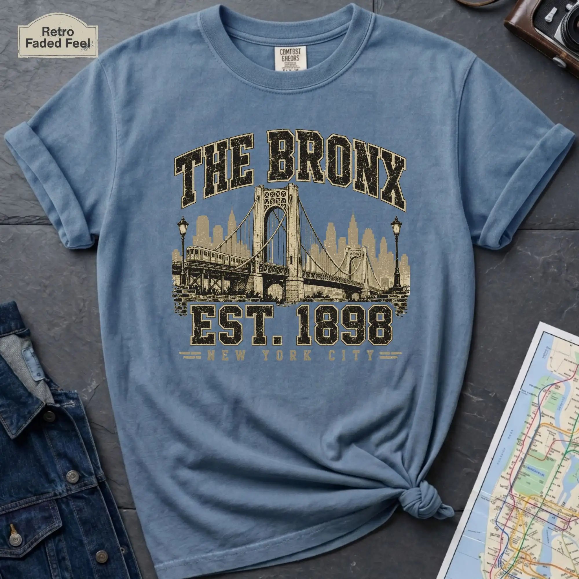 Blue t-shirt with 'The Bronx Est. 1898' graphic design on a dark surface with a map and jacket.