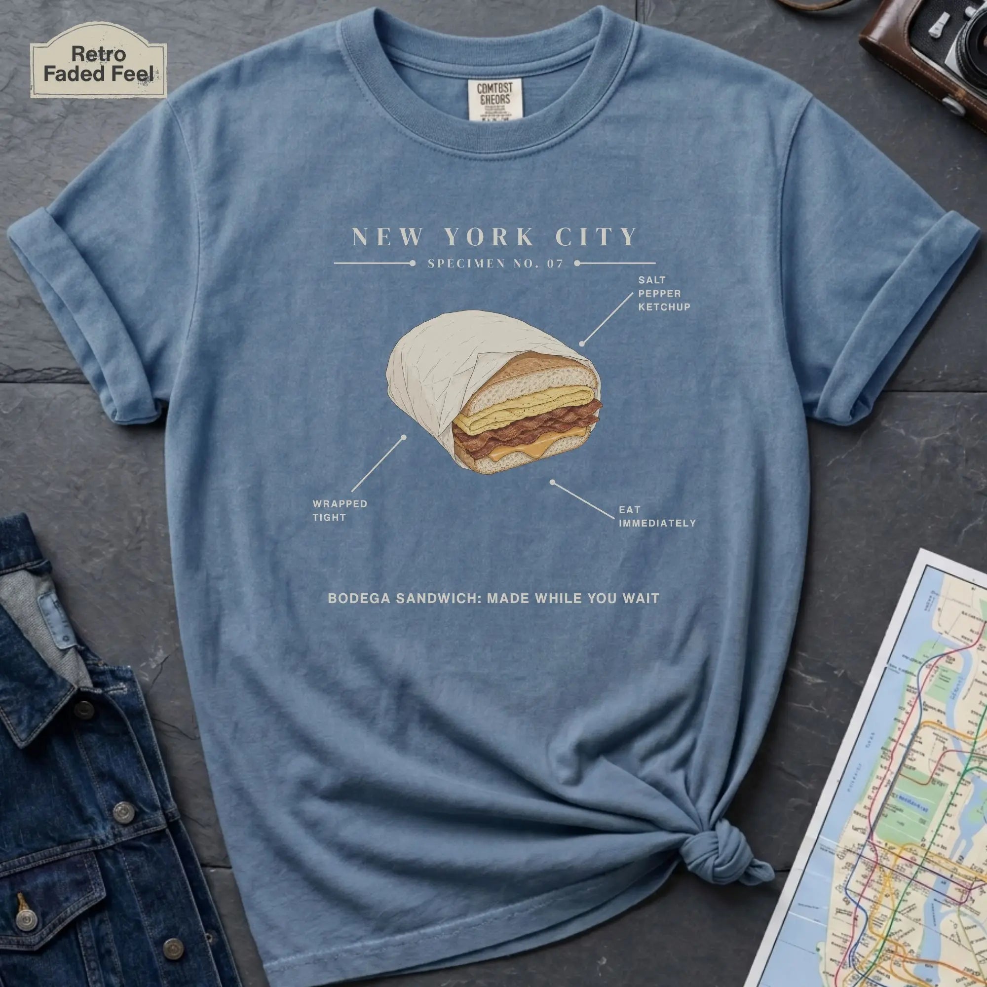 Blue t-shirt with a New York City bodega sandwich graphic on a dark surface with a map and jacket.