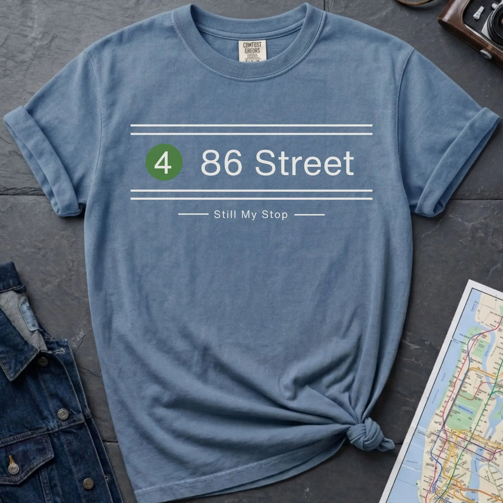 Blue t-shirt with '4 86 Street Still My Stop' text on a dark surface with jeans and a map.