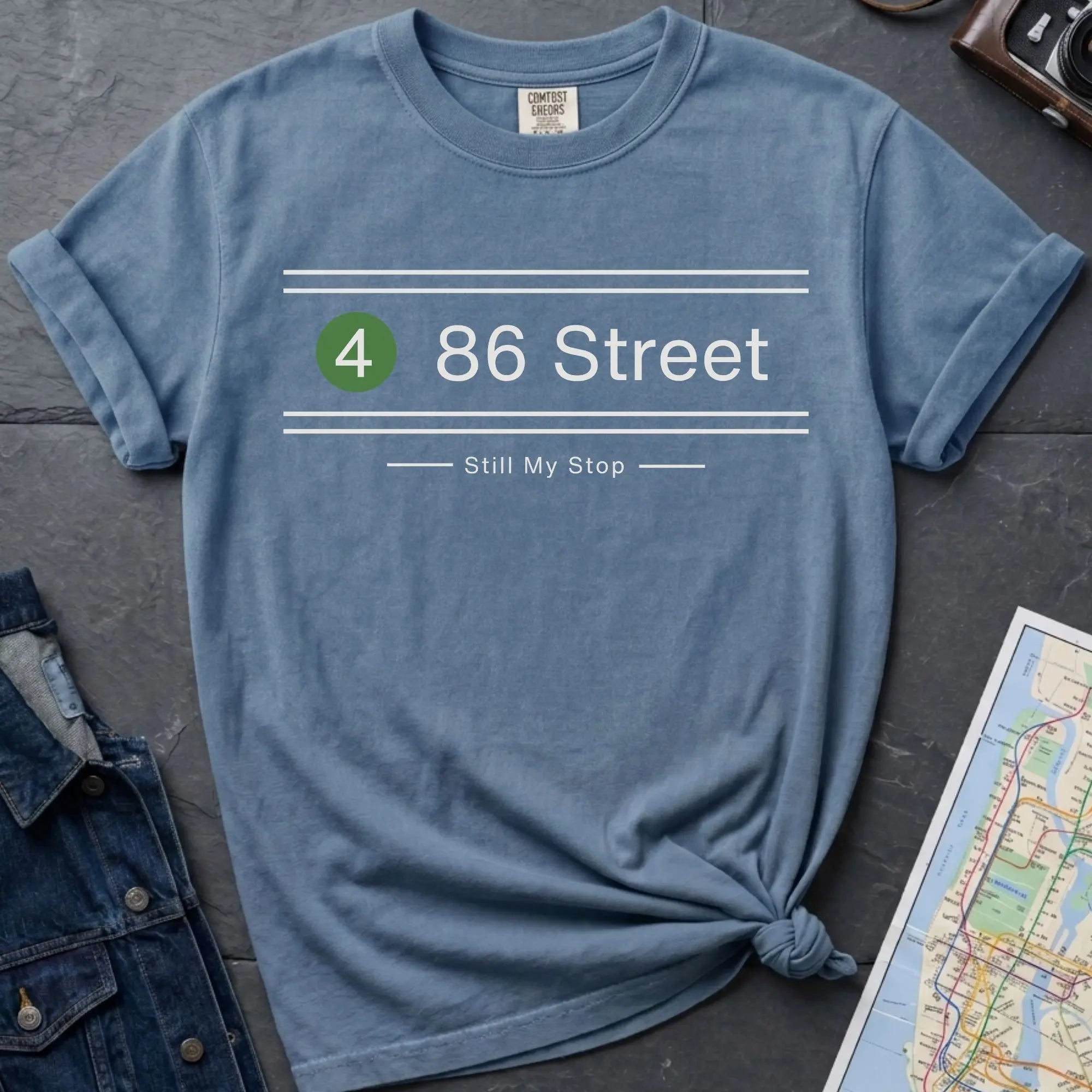 Blue t-shirt with '4 86 Street Still My Stop' text on a dark surface with jeans and a map.