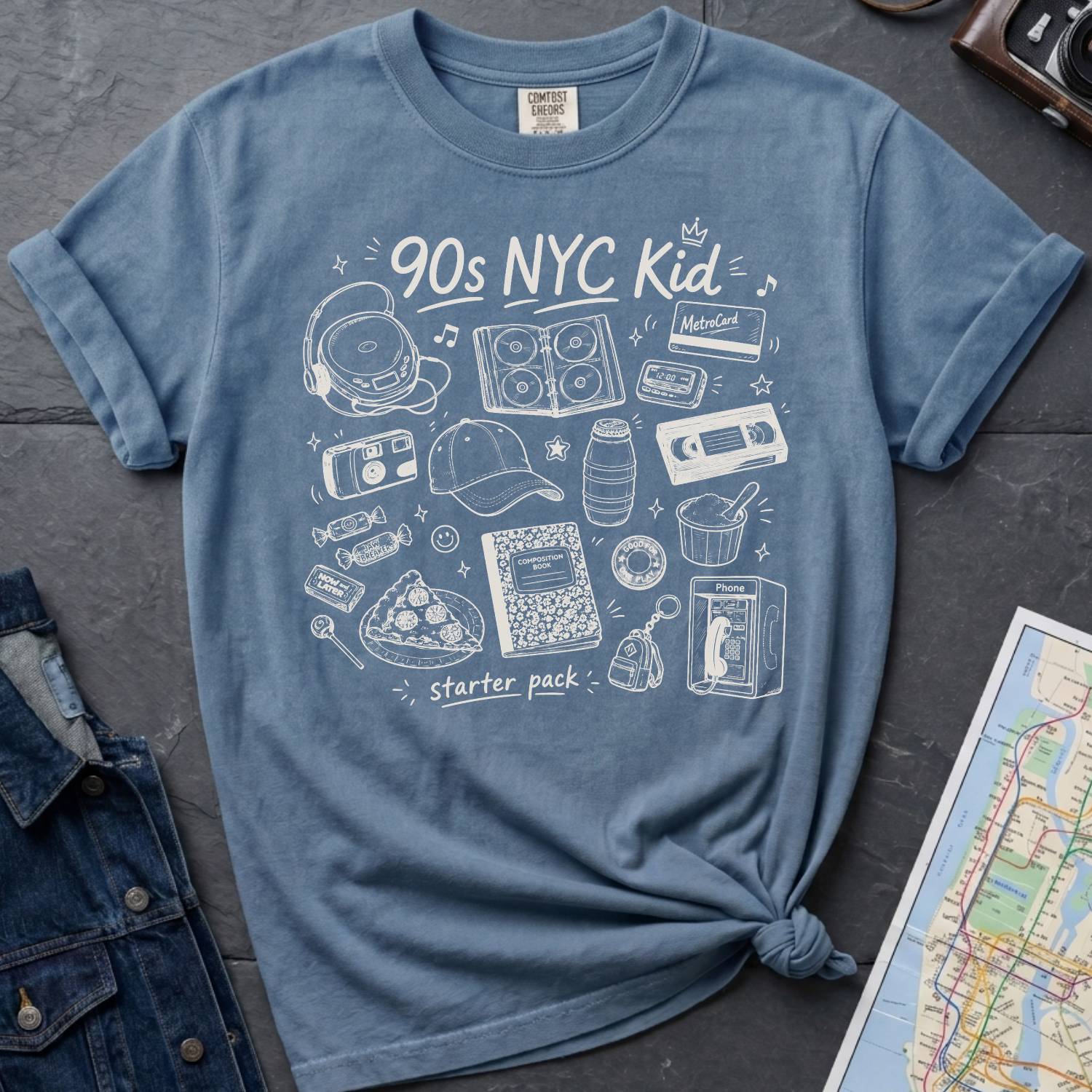 Blue Comfort Colors t-shirt with '90s NYC Kid' graphic design on a dark surface with denim jacket and map.