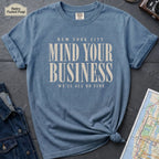 Blue t-shirt with 'New York City Mind Your Business We'll All Be Fine' text on a dark surface with a map and camera.