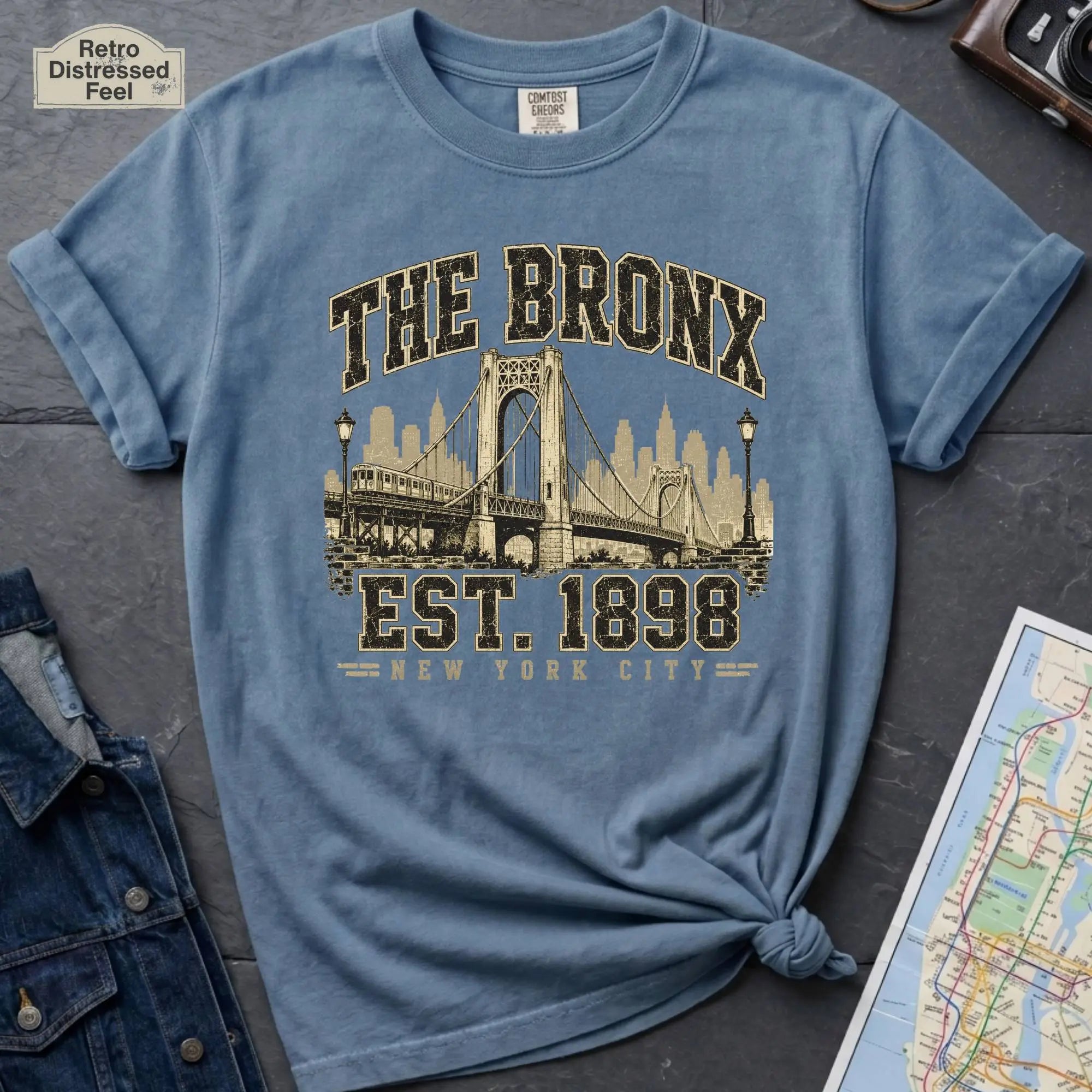 Blue t-shirt with 'The Bronx Est. 1898' design on a dark surface with a map and a denim jacket.