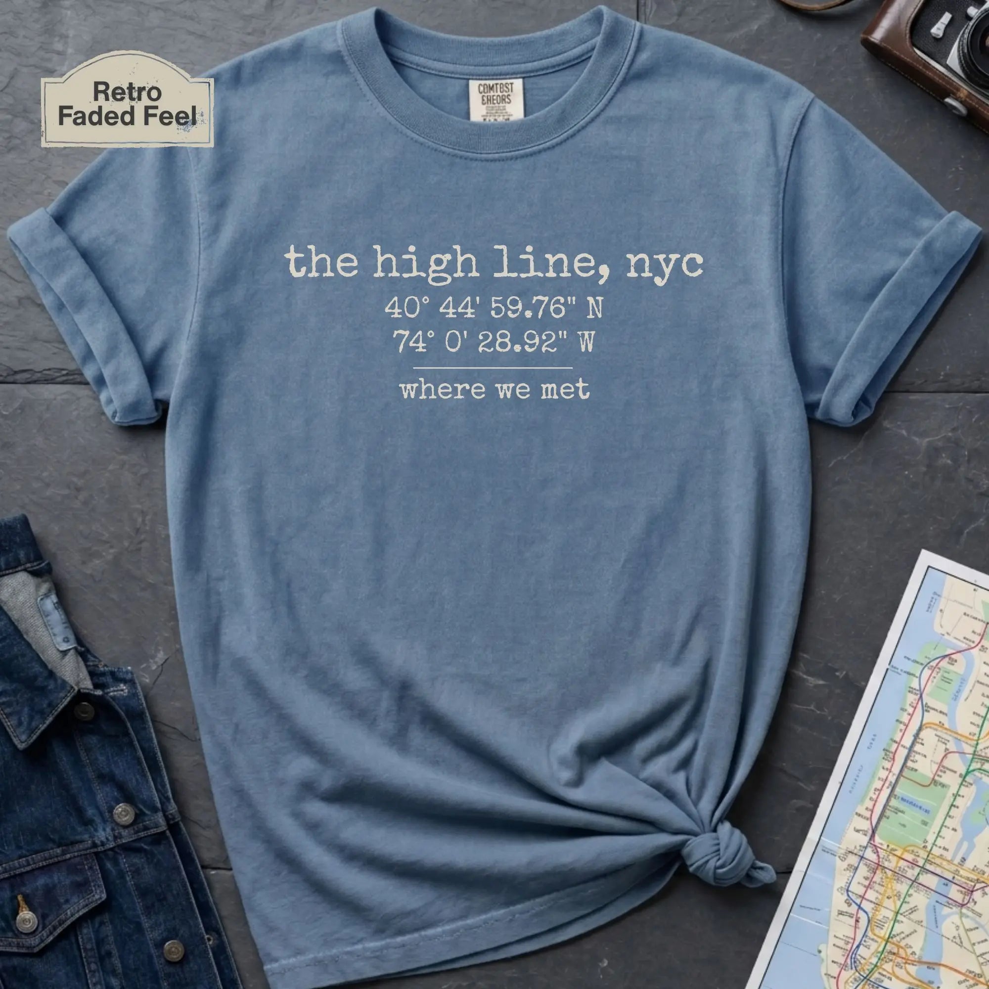 Blue t-shirt with text 'the high line, nyc' and coordinates on a dark surface with a map and jacket.