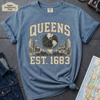 Blue t-shirt with 'Queens Est. 1683' graphic design on a dark surface with a denim jacket and a map.
