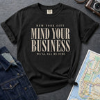 Black t-shirt with 'New York City Mind Your Business We'll All Be Fine' text on a dark surface with a map and camera.