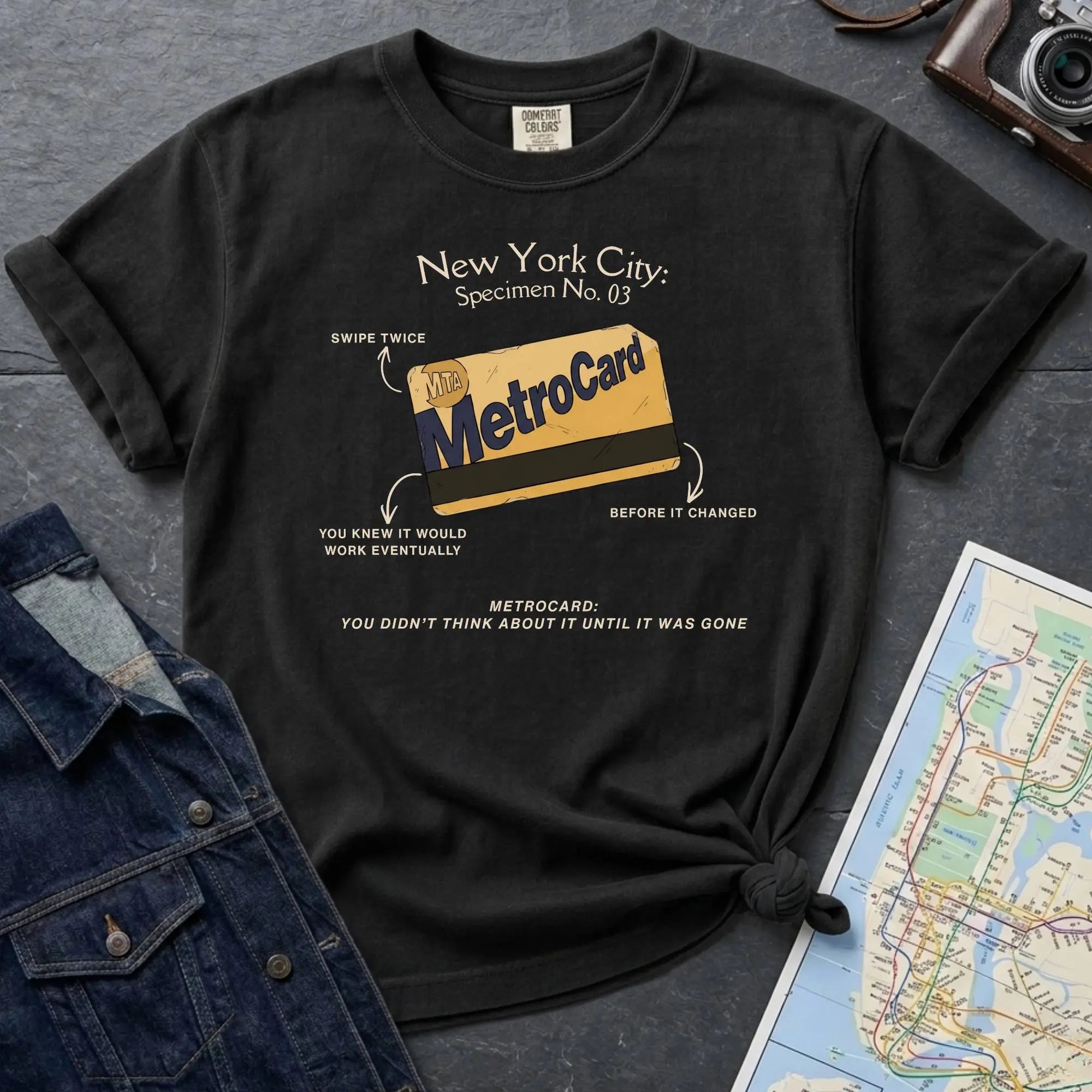 Black t-shirt with New York City MetroCard graphic on a gray surface with a map and jacket.