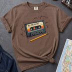 Brown t-shirt with a cassette tape graphic and pencil on a gray surface with denim jacket and map.