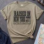 Khaki colored t-shirt with 'Raised in New York City' text on a dark surface with a jean jacket and a map.