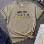 Khaki colored t-shirt with 'Queens' and subway line numbers on a dark surface with a map and camera.