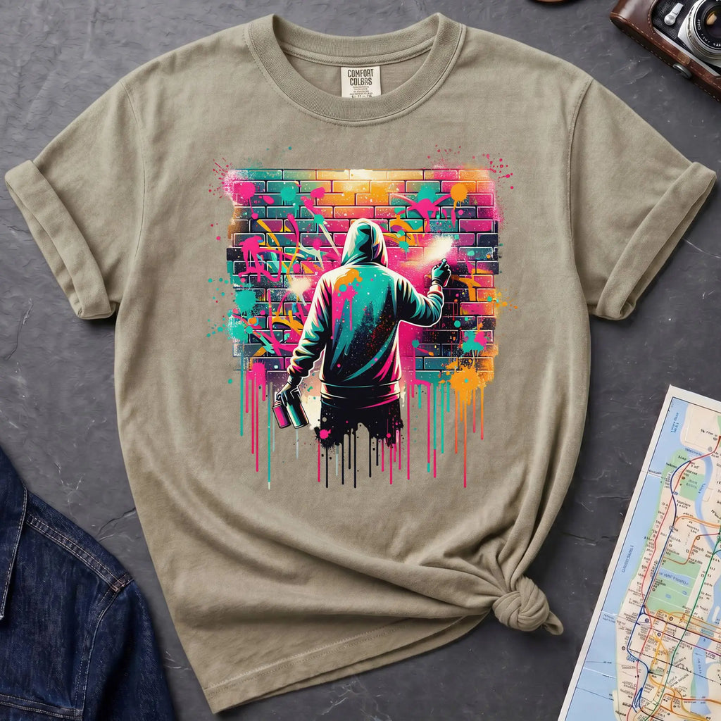 Khaki colored t-shirt with a graphic of a person spray painting a colorful mural on a brick wall.