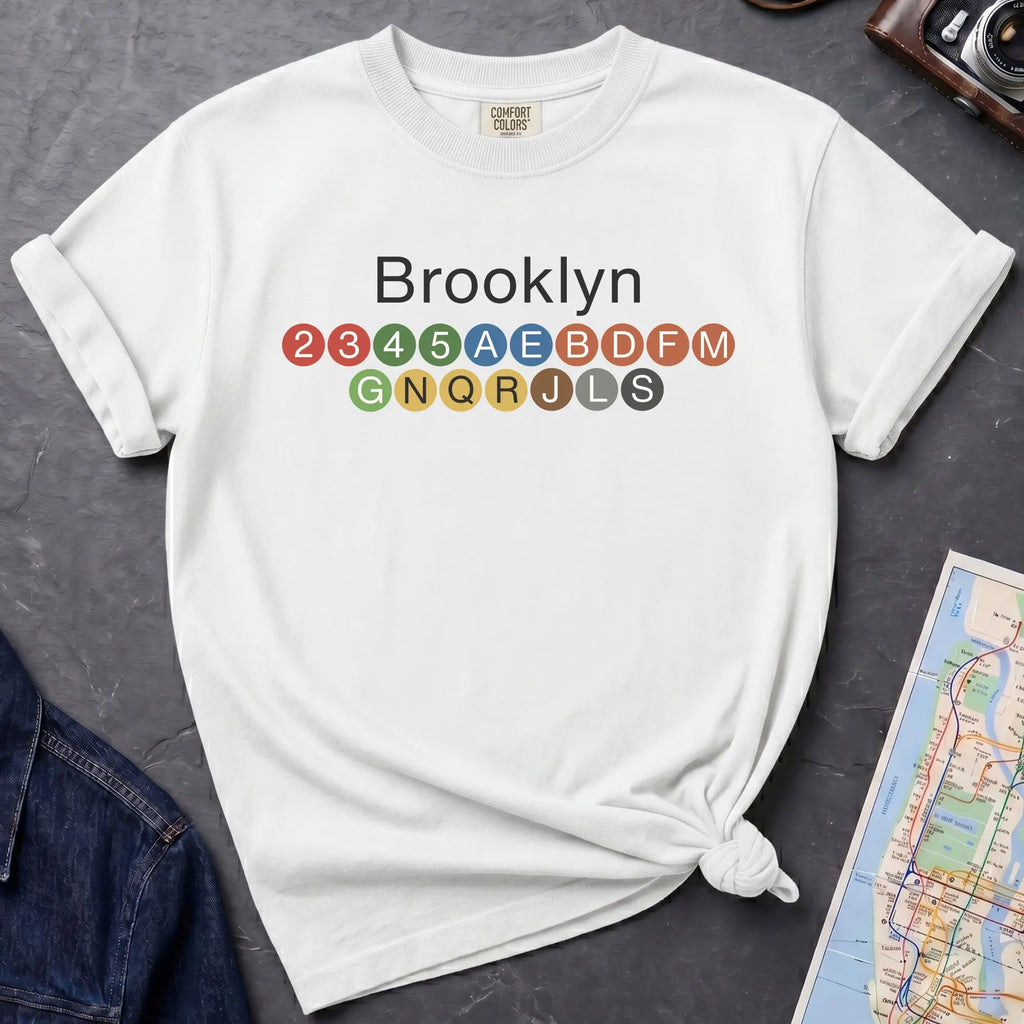 White t-shirt with 'Brooklyn' and subway line numbers on a dark surface with a map and camera.