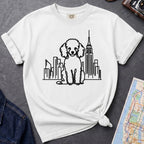 White t-shirt with a poodle and cityscape design on a dark surface with a map and camera.
