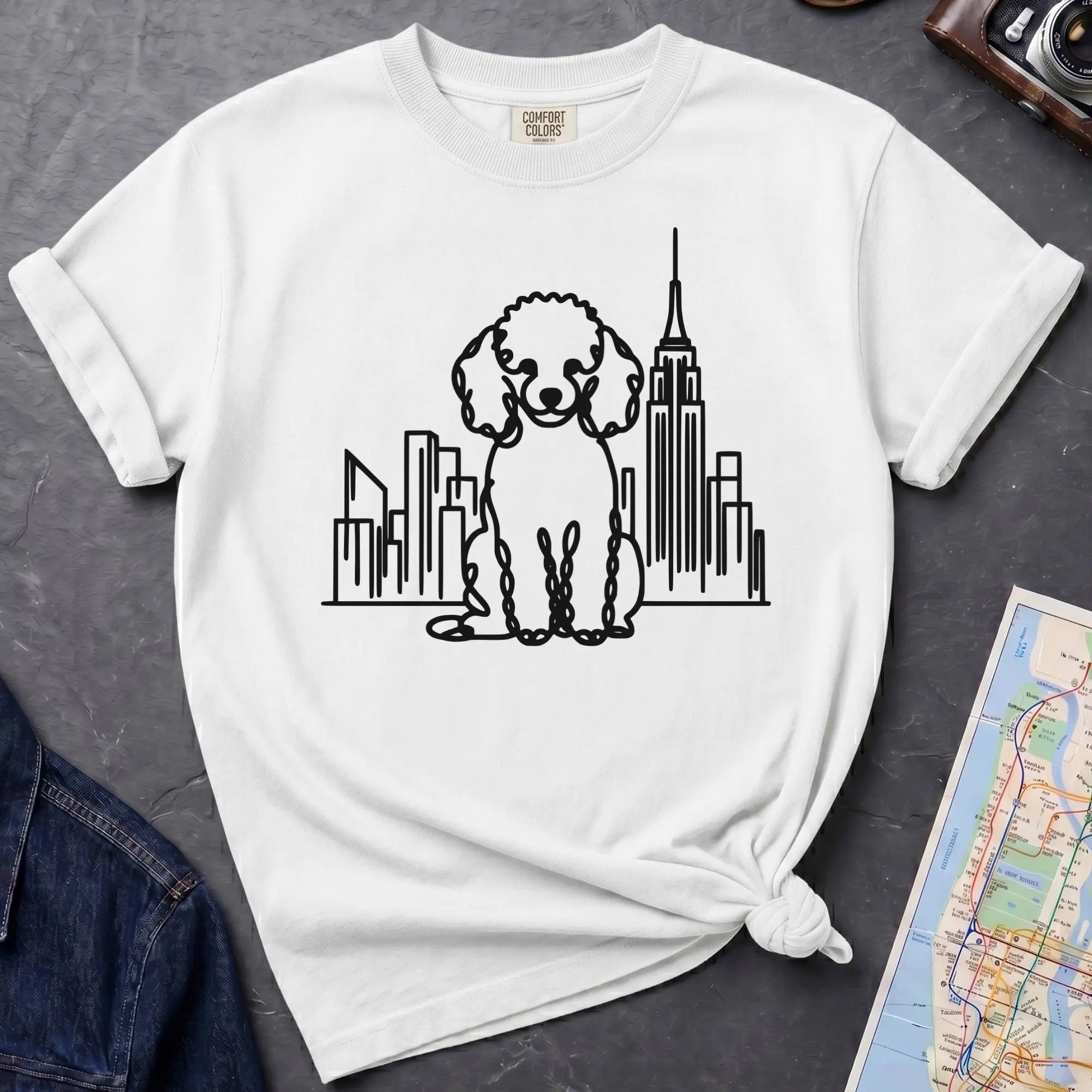 White t-shirt with a poodle and cityscape design on a dark surface with a map and camera.
