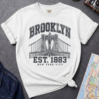 White Vintage-style Brooklyn NYC graphic t-shirt on a Bay colored Comfort Colors cotton shirt, featuring the Brooklyn Bridge and “Brooklyn Est. 1883” text.