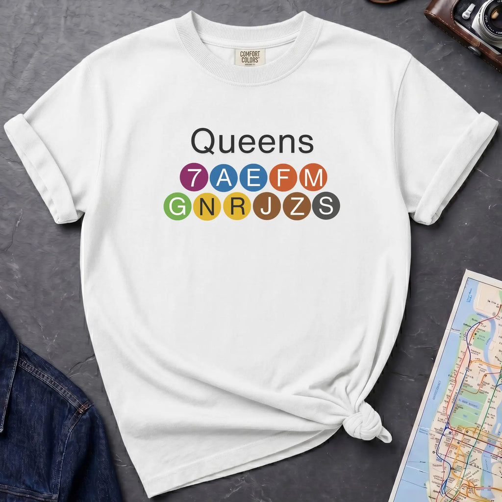 White t-shirt with 'Queens' and NYC subway lines on a dark surface with a map and denim jacket.