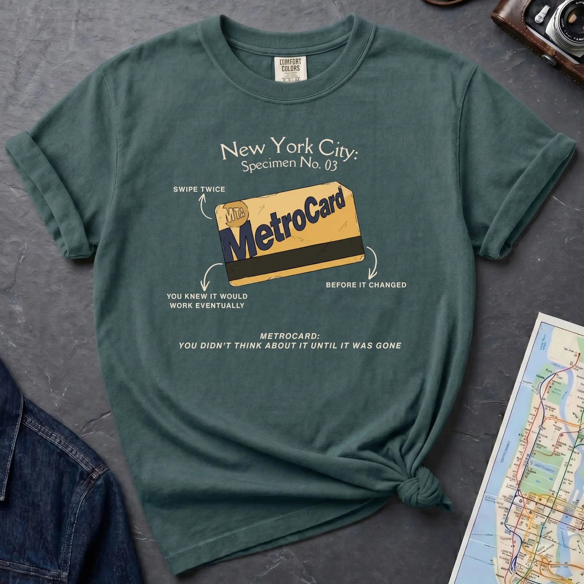 Blue spruce t-shirt with New York City MetroCard graphic on a gray surface with a map and jacket.