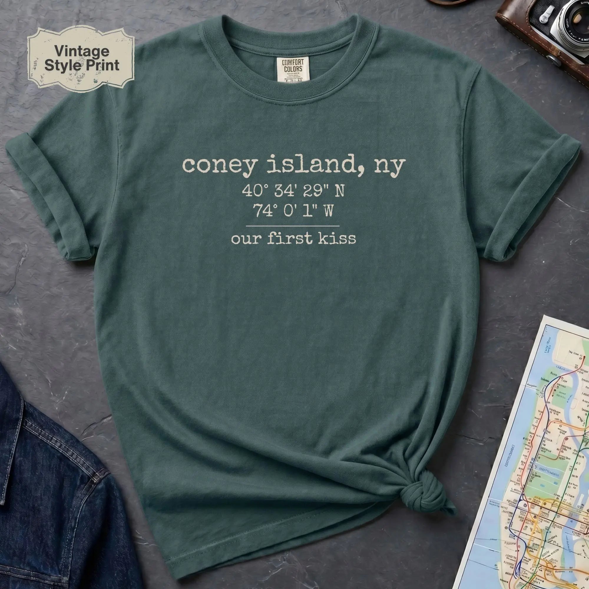 Blue Spruce t-shirt with text 'coney island, ny' and coordinates on a gray surface with a map and camera.