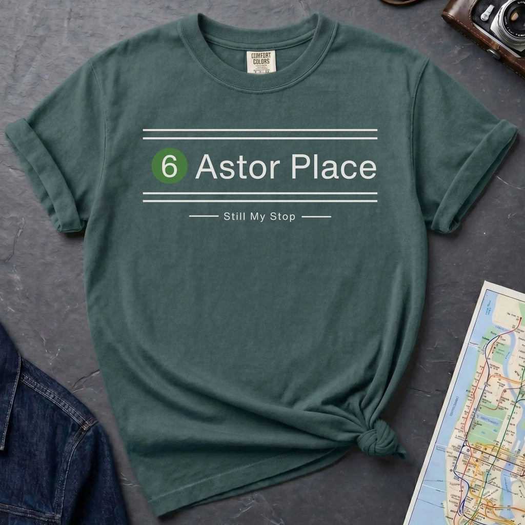 Blue Spruce t-shirt with '6 Astor Place Still My Stop' design on a dark surface with a map and jeans.