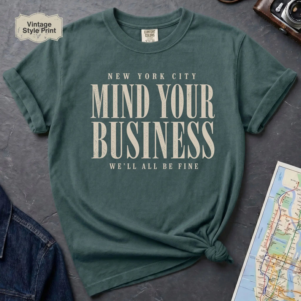 Blue Spruce t-shirt with 'New York City Mind Your Business We'll All Be Fine' text on a dark surface with a map and camera.