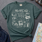 Blue Spruce Comfort Colors t-shirt with '90s NYC Kid' graphic design on a gray surface with a map and a denim jacket.