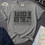 Granite colored t-shirt with 'Raised in New York City' text on a dark surface with a jean jacket and a map.