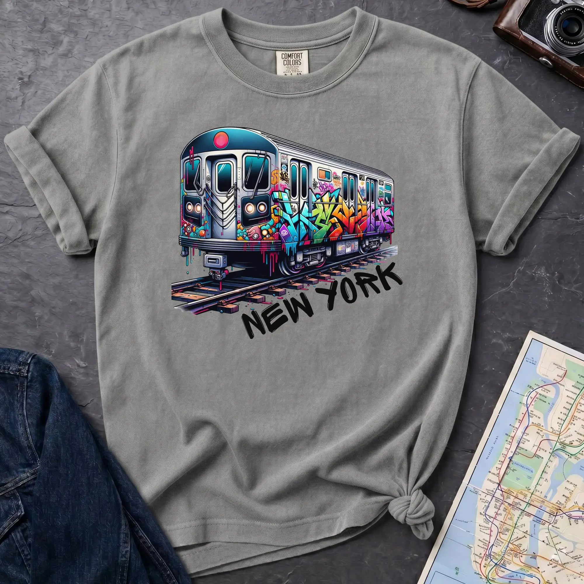 Granite colored t-shirt with a colorful train graphic and 'New York' text on a gray surface with a map and denim jacket.