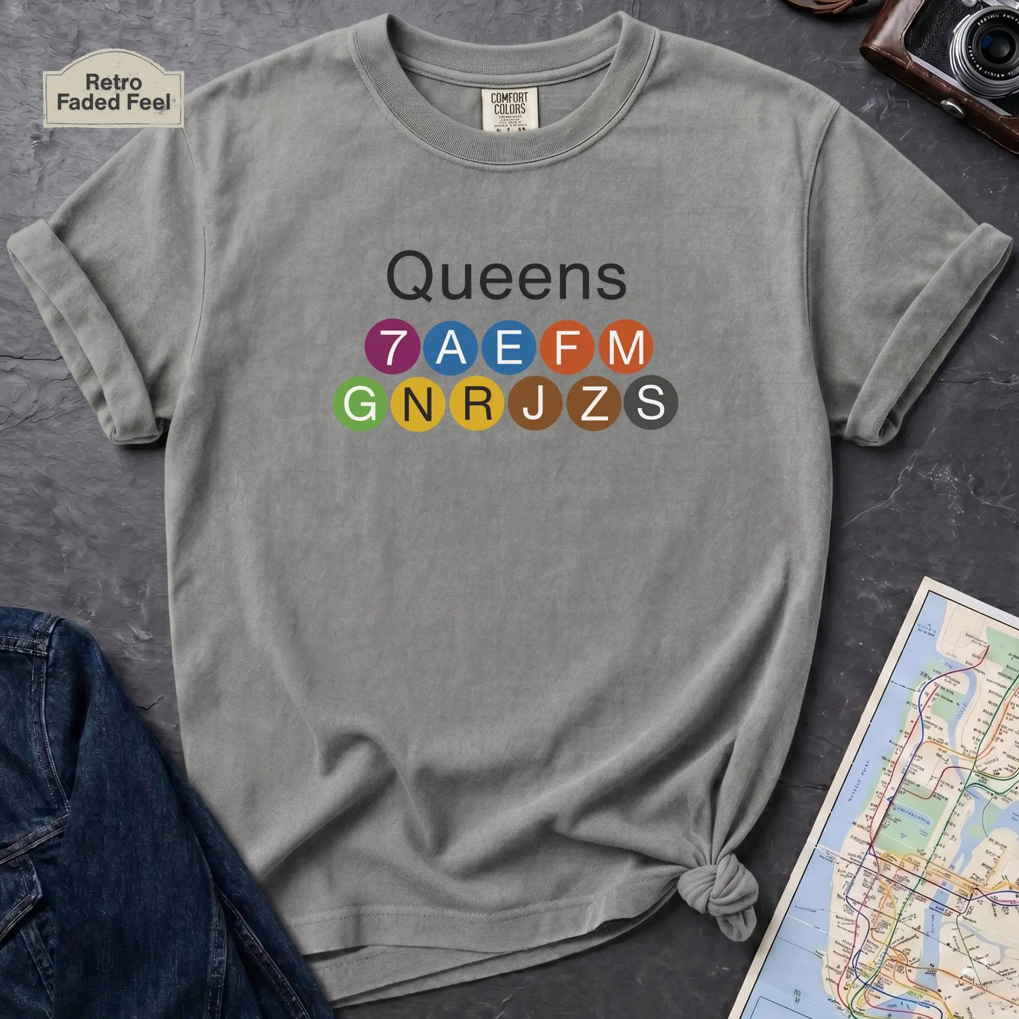 Granite colored t-shirt with 'Queens' and NYC subway lines on a dark surface with a map and denim jacket.