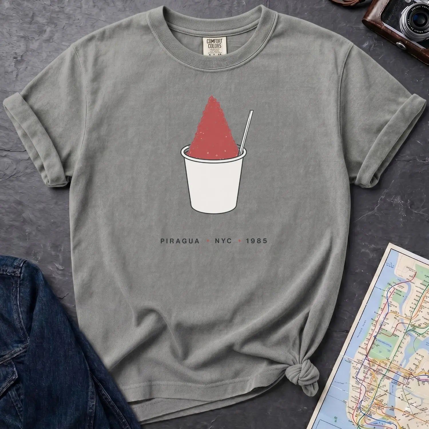 Gray Comfort Colors t-shirt with a piragua graphic and text, placed on a dark surface with a map and denim jacket.