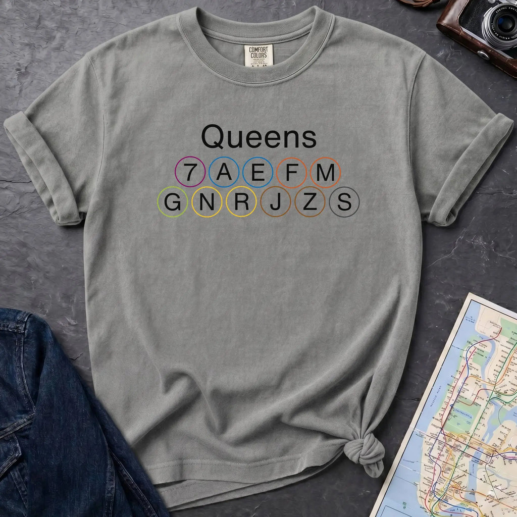 Granite colored t-shirt with 'Queens' and subway line numbers on a dark surface with a map and camera.