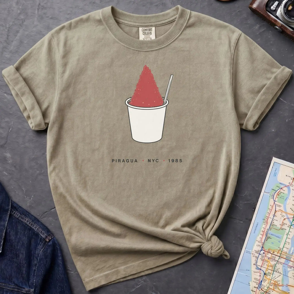 Khaki colored Comfort Colors t-shirt with a piragua graphic and text, placed on a dark surface with a map and denim jacket.