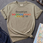 Khaki colored t-shirt with 'Brooklyn' and subway line numbers on a dark surface with a map and camera.