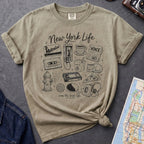 Khaki colored t-shirt with New York-themed graphics on a gray surface with a map and camera.
