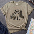 Khaki colored t-shirt with a poodle and cityscape design on a dark surface with a map and camera.