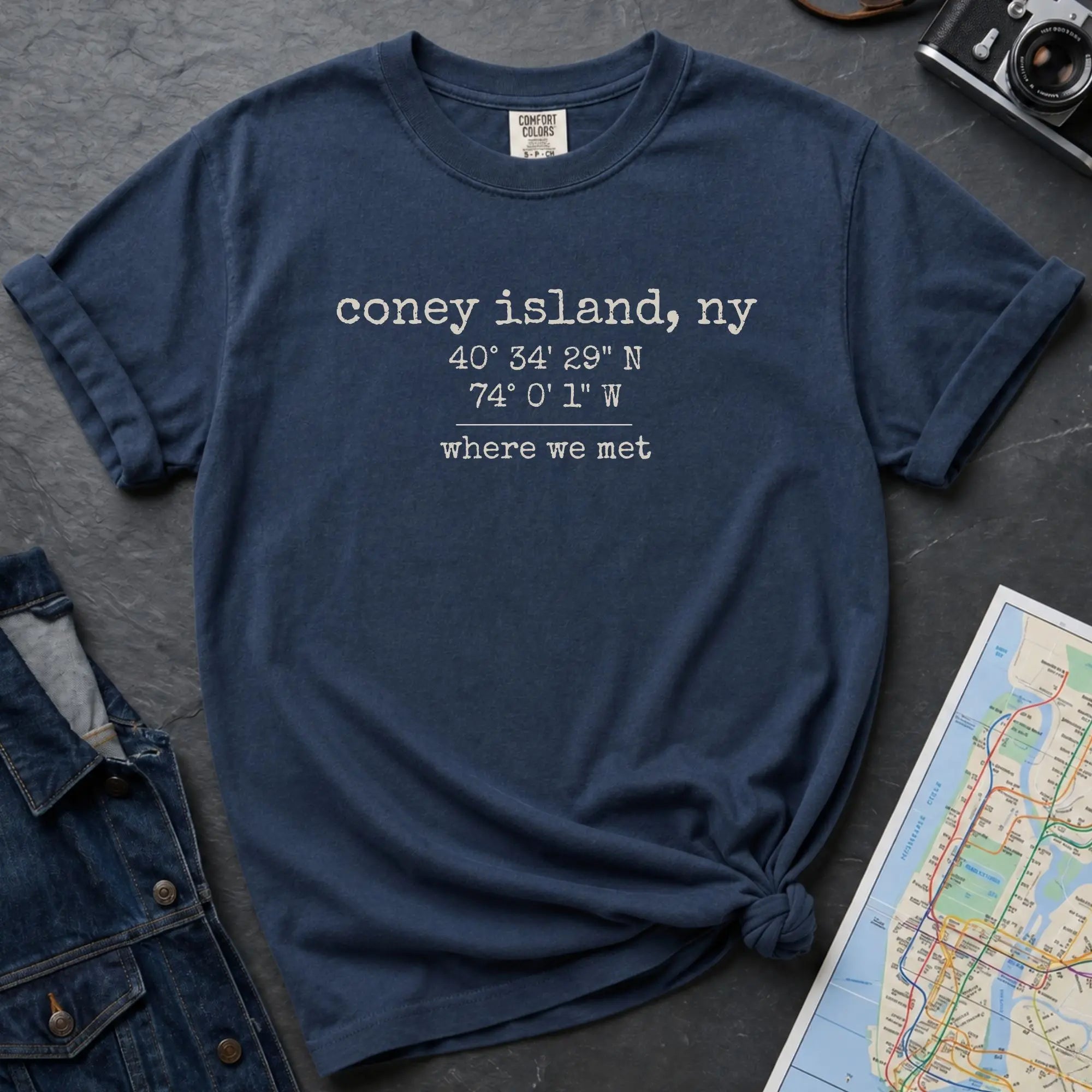Blue t-shirt with text 'coney island, ny 40°34'29"N 74°0'1"W where we met' on a dark surface with a map and denim jacket.