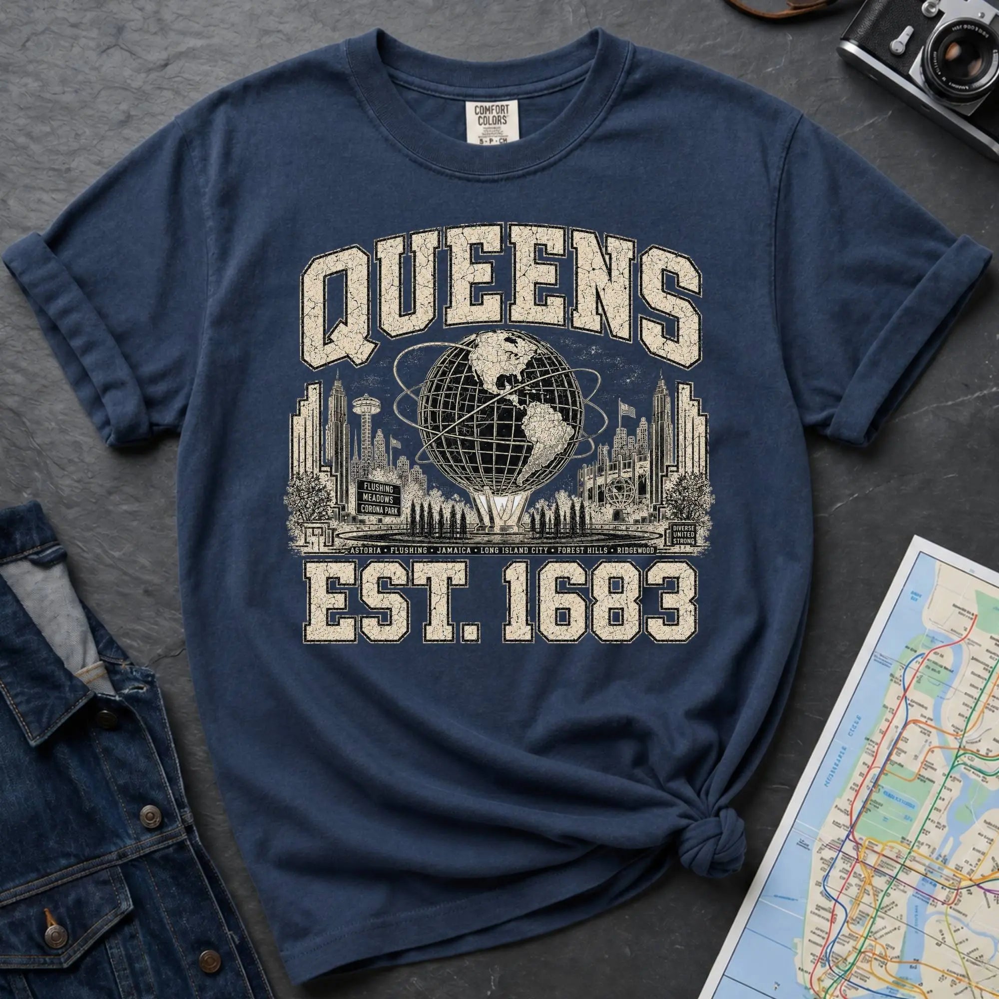 Navy t-shirt with 'Queens Est. 1683' graphic design on a dark surface with a denim jacket and a map.