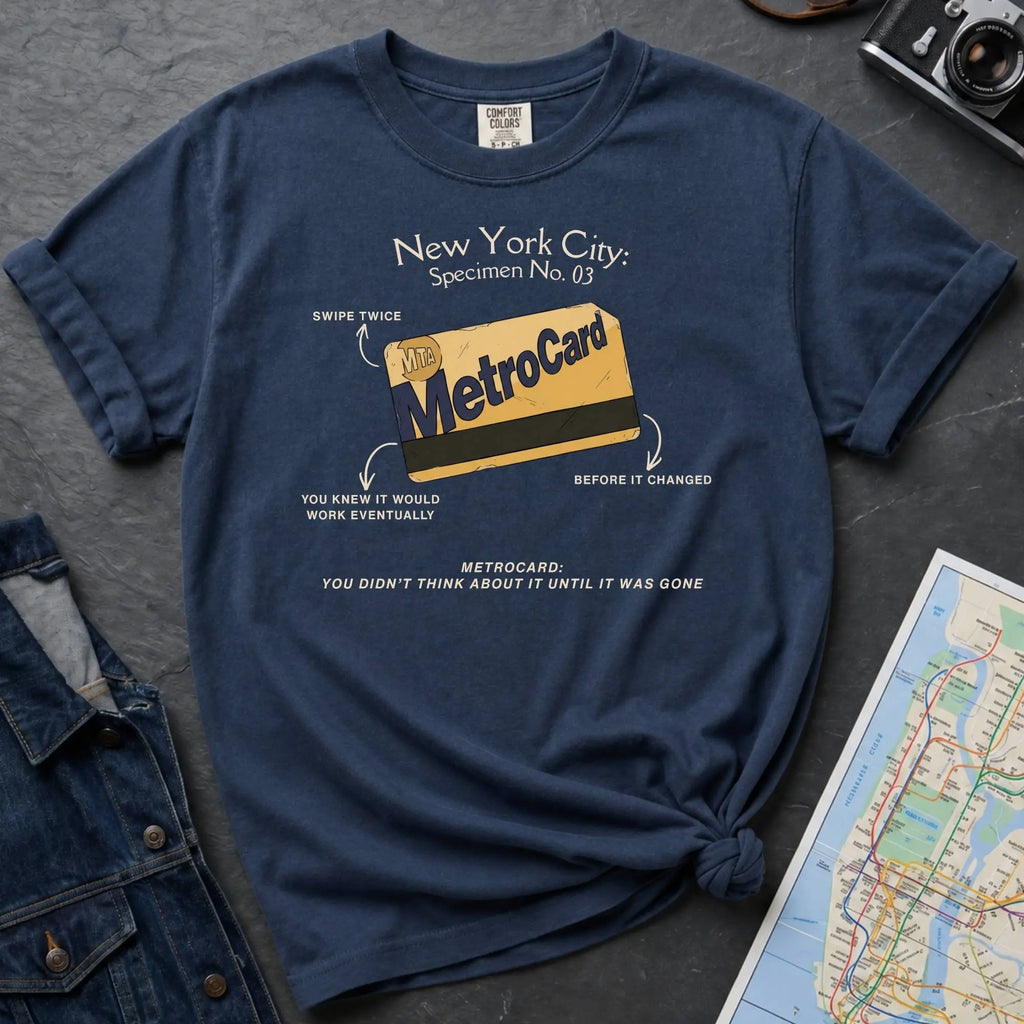 Navy t-shirt with New York City MetroCard graphic on a gray surface with a map and jacket.